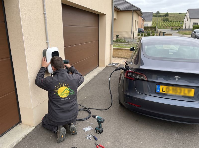Électricien Ecolive Lux fixant une borne de recharge murale à côté d'un garage, voiture électrique branchée